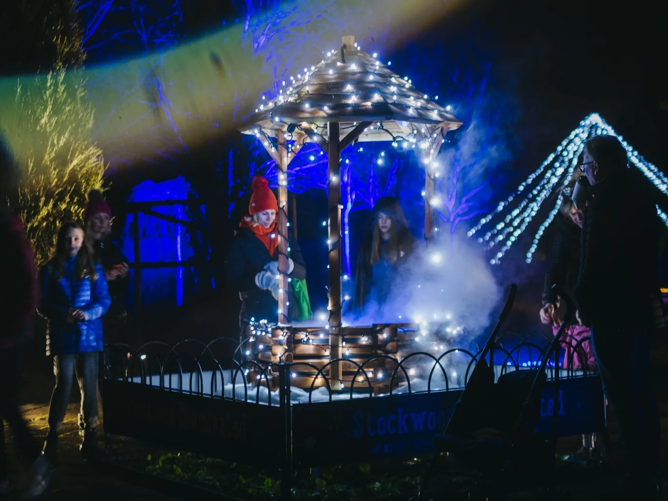 A wooden gazebo wrapped in fairy lights with mist rising from it, visitors standing around